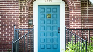 A blue door with a numeric address sign above it, set in a brick building with a small porch and a metal railing.