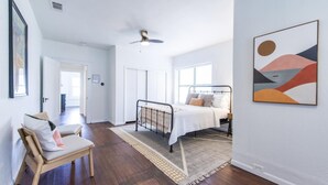 A bedroom with a bed, a chair, a ceiling fan, and a painting on the wall.