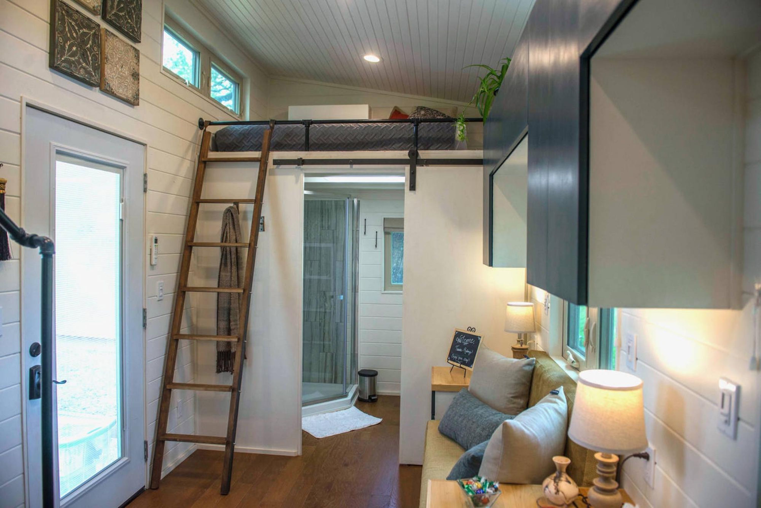 A loft bedroom with a bunk bed, a ladder, a bathroom with a shower, and a living area with a couch and lamp.