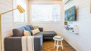 A cozy living room with a grey sectional sofa, a small white stool, a gold floor lamp, a TV mounted on the wall, and a window with blinds. - Pecan Grove Container Tiny Home Country Setting 12 min to Baylor Magnolia (Waco)