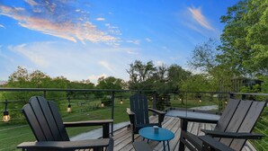 A wooden deck with Adirondack chairs and a small table, overlooking a grassy area with trees and a clear sky. - Pecan Grove Container Tiny Home Country Setting 12 min to Baylor Magnolia (Waco)