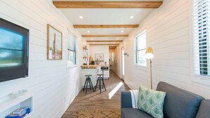 A narrow hallway with a kitchen area at the end, wooden beams on the ceiling, and a sofa with a patterned pillow. - Pecan Grove Container Tiny Home Country Setting 12 min to Baylor Magnolia (Waco)
