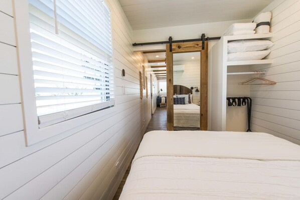 A narrow hallway leading to a bedroom with a bed, a wooden bench, and a clothing rack. - The Meadows Container Home in Country Setting (Waco)