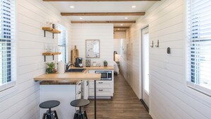 A narrow kitchen with white paneling, wooden beams, and a central island with stools. - The Meadows Container Home in Country Setting (Waco)