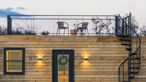 A wooden building with a balcony, two chairs, a door with a wreath, and a spiral staircase. - The Meadows Container Home in Country Setting (Waco)