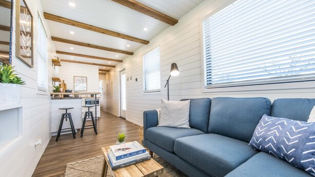 A modern living room with a blue sofa, wooden beams on the ceiling, and a kitchen area in the background. - The Meadows Container Home in Country Setting (Waco)