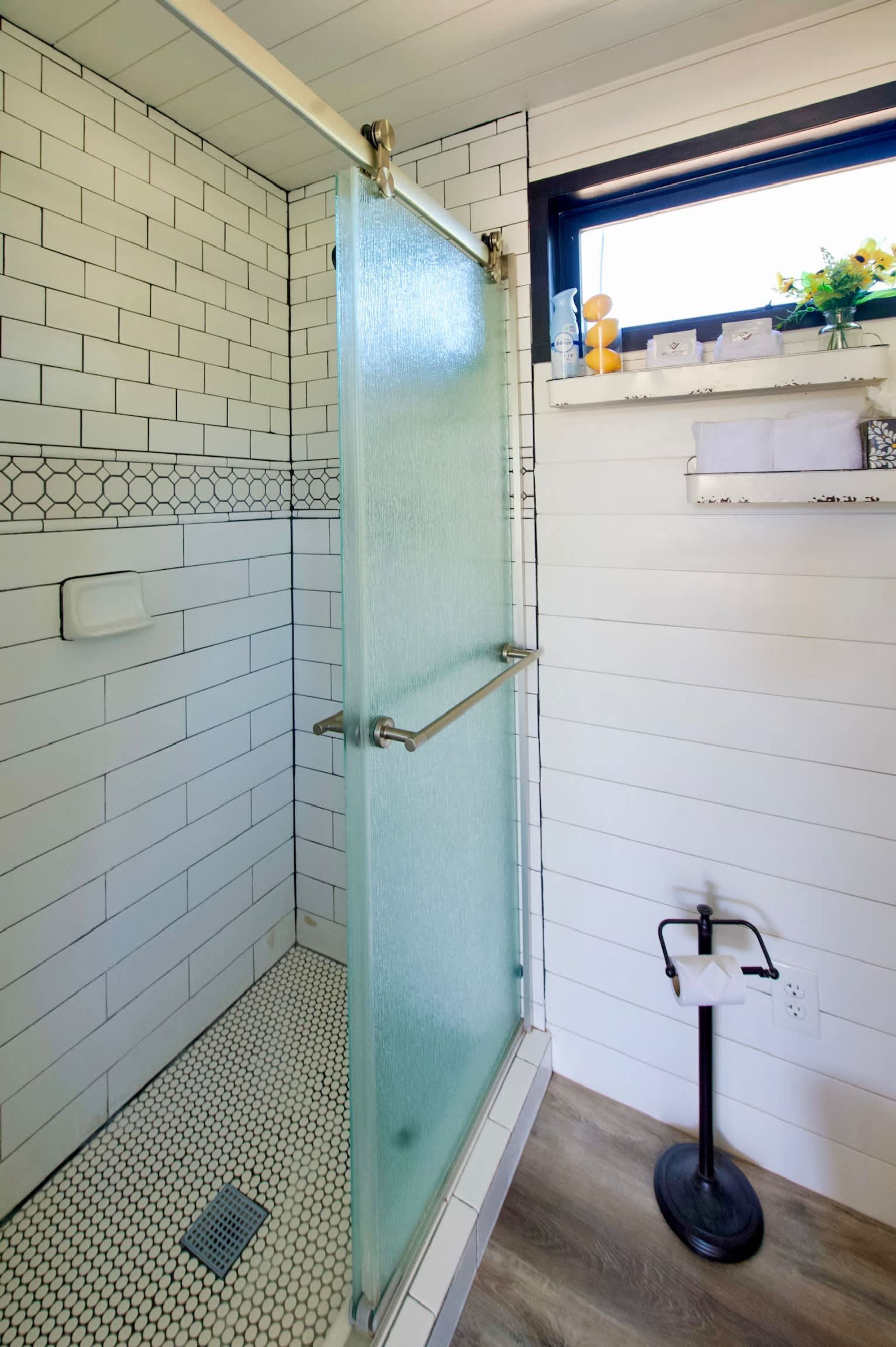 Zona de ducha con puerta de vidrio, paredes de azulejos blancos, suelo de madera y una ventana con repisa superior.