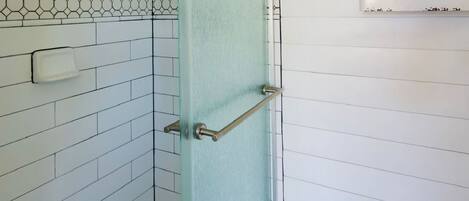 Zona de ducha con puerta de vidrio, paredes de azulejos blancos, suelo de madera y una ventana con repisa superior.