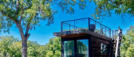 Une cabane en bois dotée d’une structure vitrée, située dans une zone herbeuse bordée d’arbres.