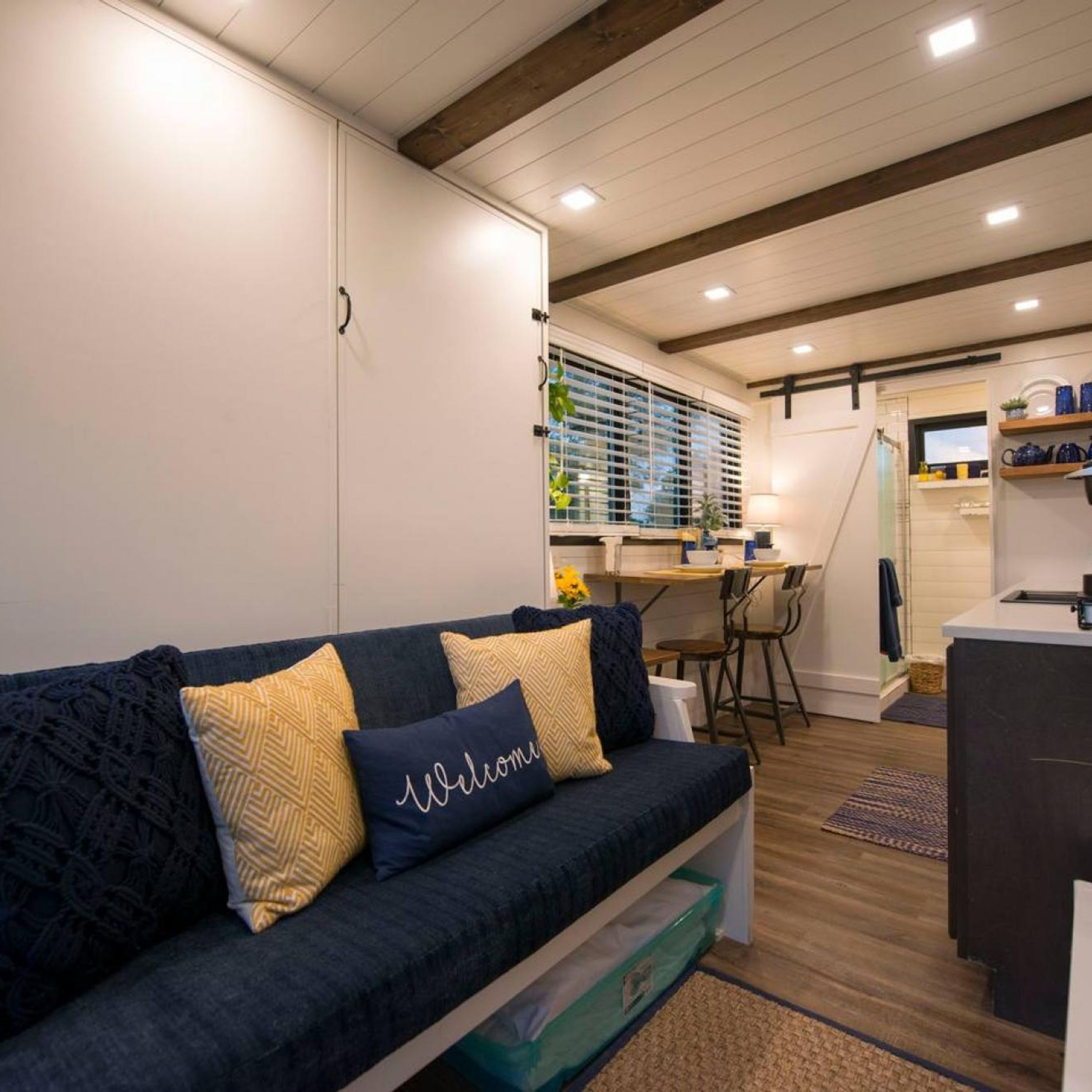 A cozy living room with a dark blue sofa, a kitchen area with wooden flooring, and a dining table.