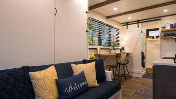 A cozy living room with a dark blue sofa, a kitchen area with wooden flooring, and a dining table.
