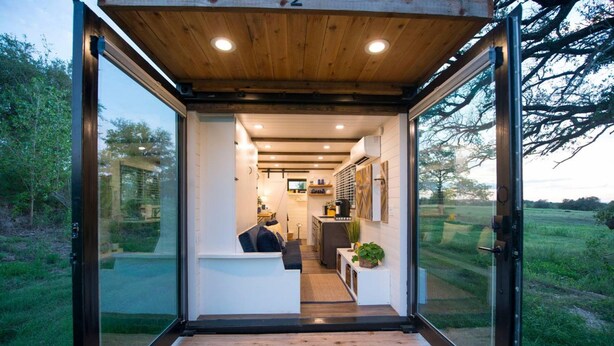 A modern, open-plan living space with a wooden ceiling and large glass doors leading to a garden. - Yellow Blue Elegant Container Tiny House (Waco)