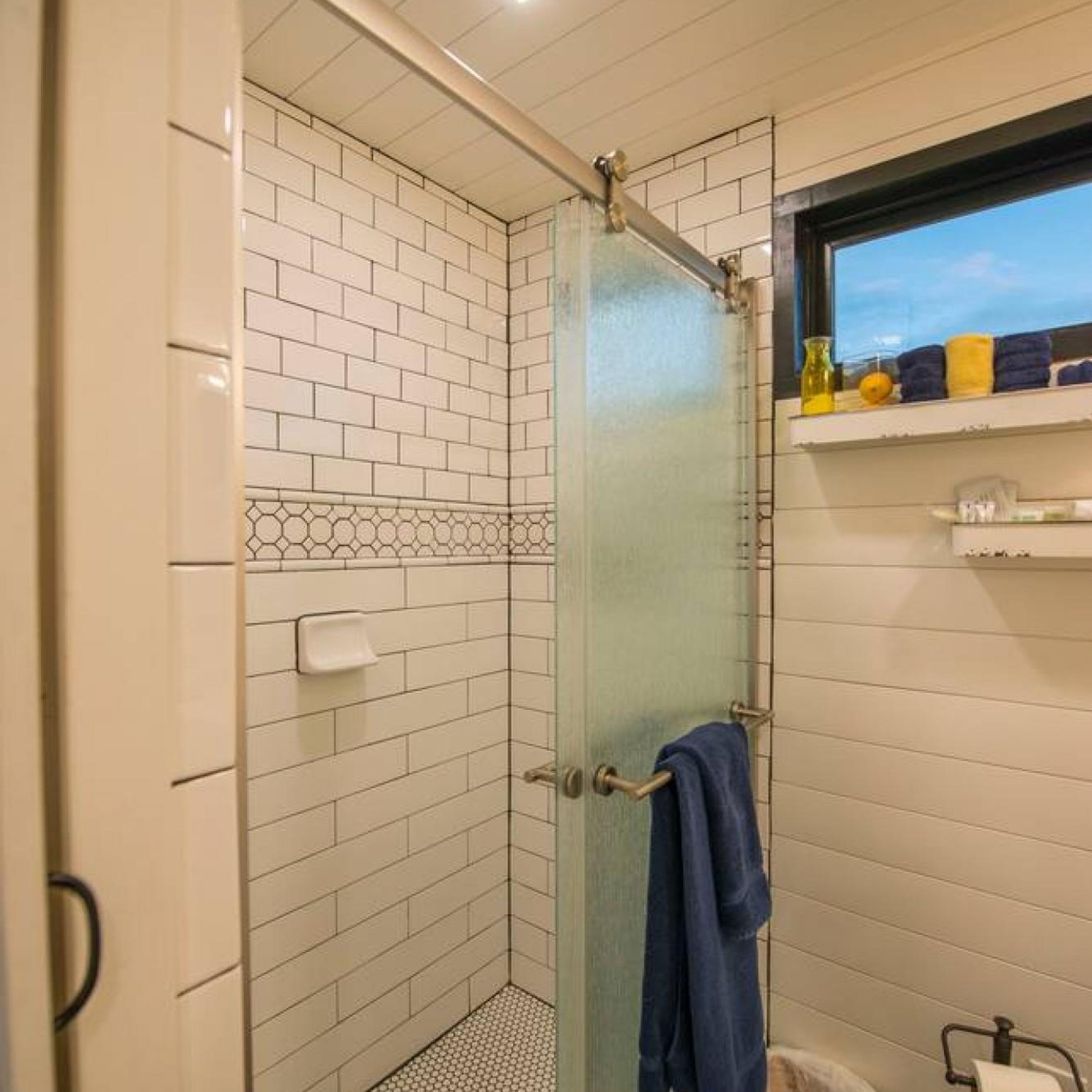 A small, enclosed shower area with a glass door, white tiled walls, and a window above the sink.