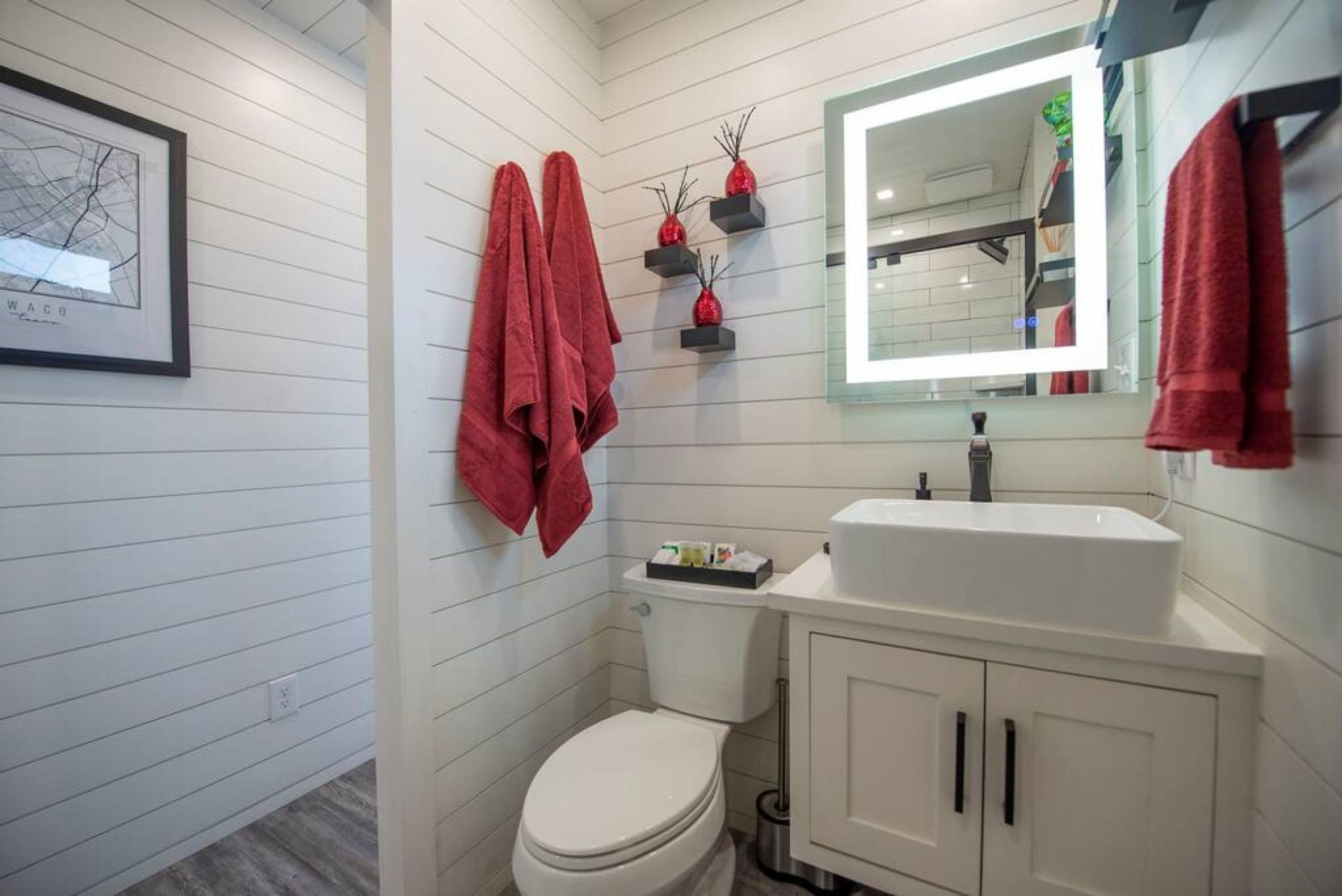 Une salle de bain avec des toilettes blanches, un lavabo blanc et un miroir avec un éclairage intégré. Des serviettes rouges sont accrochées au mur, et une carte encadrée est posée sur le mur, à gauche.