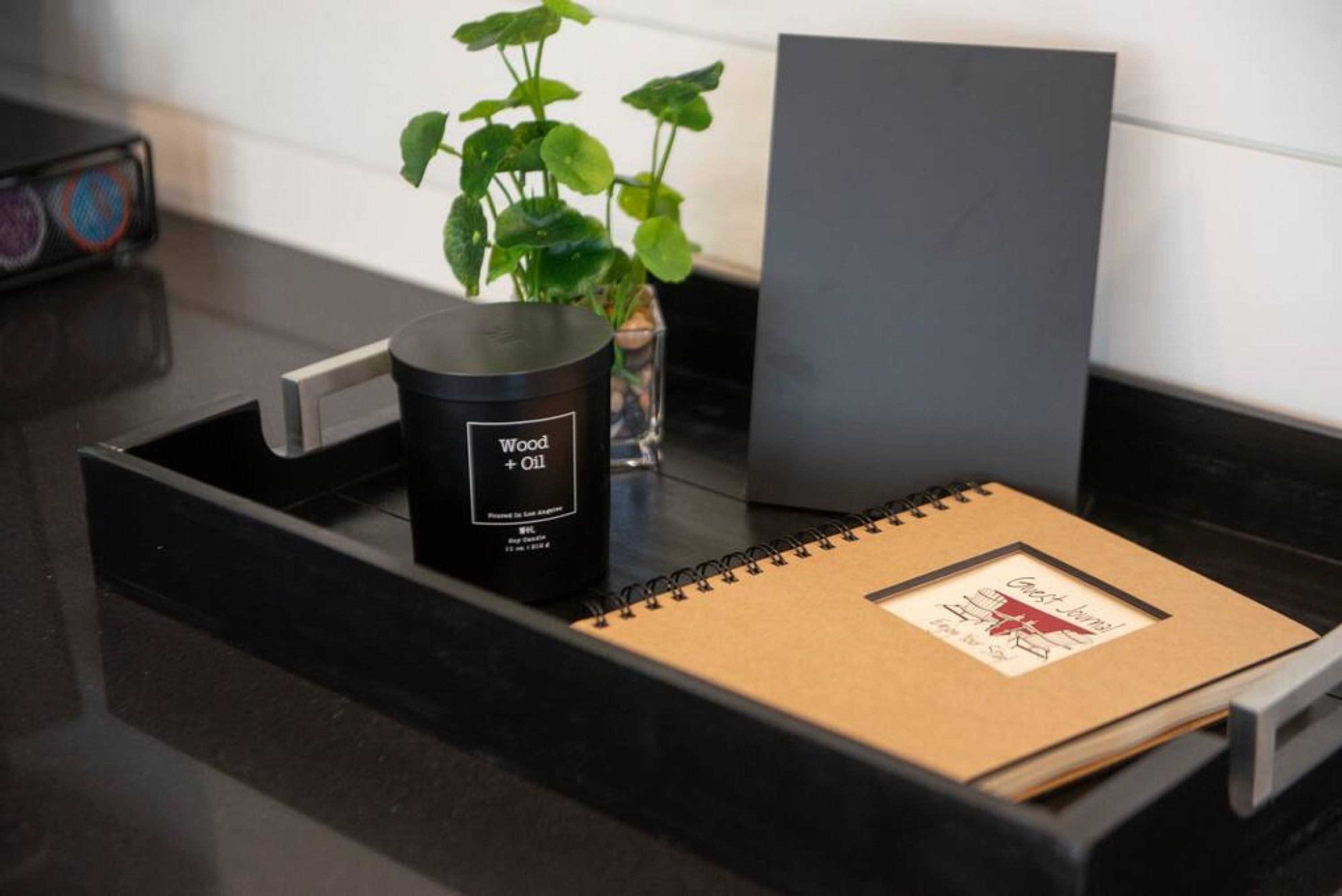 A black tray with a notebook, a candle labeled "Wood + Oil," and a potted plant on a kitchen counter.