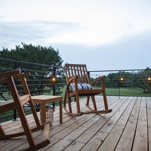 A wooden deck with two rocking chairs and a table, overlooking a grassy area with trees. - The Bluebonnet- Container Home Country Setting Near Waco Attractions (Waco)
