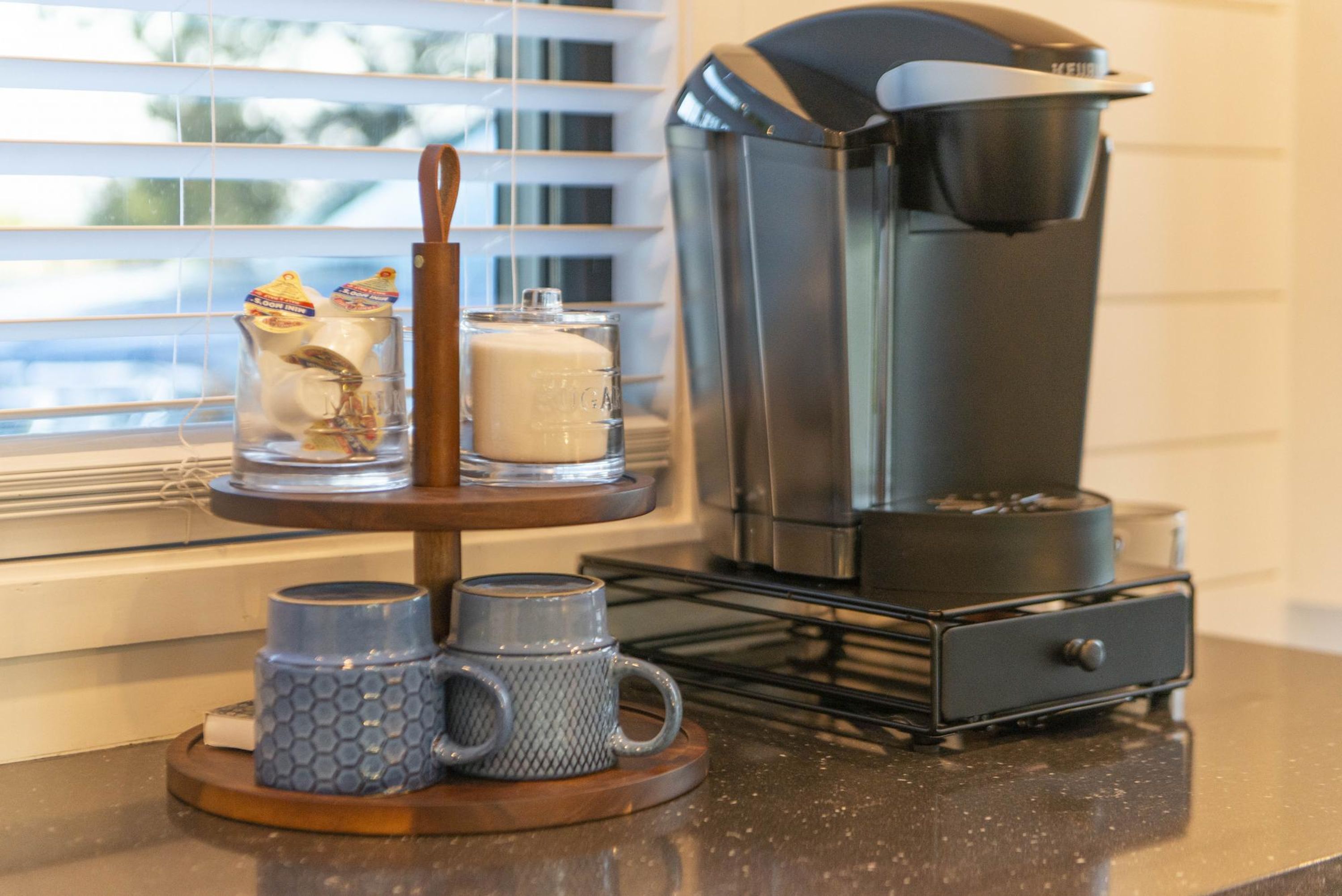 A coffee maker on a countertop with two mugs and a glass jar.