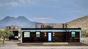 A modern log cabin with a blue door, a small porch with chairs, and a view of mountains in the background. - New Longhorn Stunning Container Home-in Alpine (Alpine)
