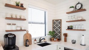 A modern kitchen with white cabinetry, a coffee maker, a sink, a microwave, and various decorative items on shelves. - New Longhorn Stunning Container Home-in Alpine (Alpine)