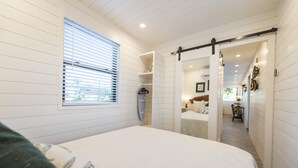 A bedroom with a bed, a window with blinds, a sliding door leading to a bathroom, and a hallway with a table and chairs. - The Woods- Container Tiny House 12 min to Magnolia Baylor Downtown (Waco)
