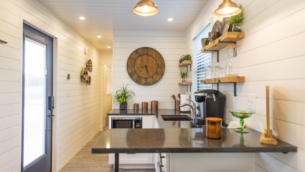 A narrow kitchen with a wooden wall clock, pendant lights, and a small dining area. - The Woods- Container Tiny House 12 min to Magnolia Baylor Downtown (Waco)