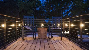 A wooden deck with two rocking chairs and a small table, surrounded by a metal railing and string lights. - The Woods- Container Tiny House 12 min to Magnolia Baylor Downtown (Waco)