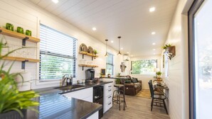 A modern kitchen with a central island, built-in shelves, and a dining area with a sofa and chairs. - The Woods- Container Tiny House 12 min to Magnolia Baylor Downtown (Waco)