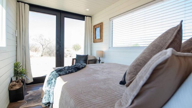 A bedroom with a large bed, a window with blinds, a bedside table with a lamp, and a view of the outdoors. - New The Cowboy Boots-shipping Container Home (Alpine)