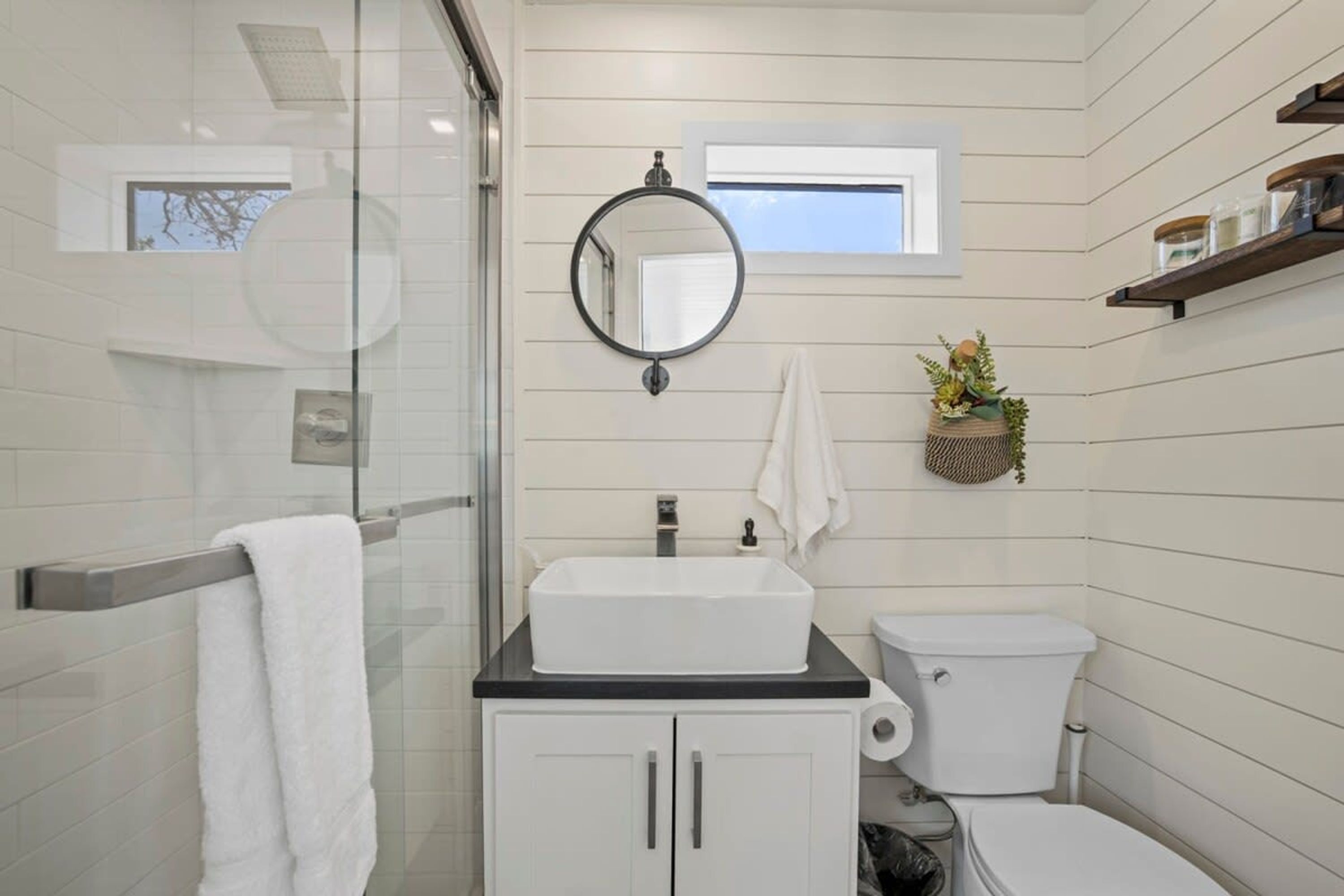 Een badkamer met een douchecabine van glas, een witte wastafel met een zwart aanrecht, een wit toilet en een klein raam boven de spiegel.