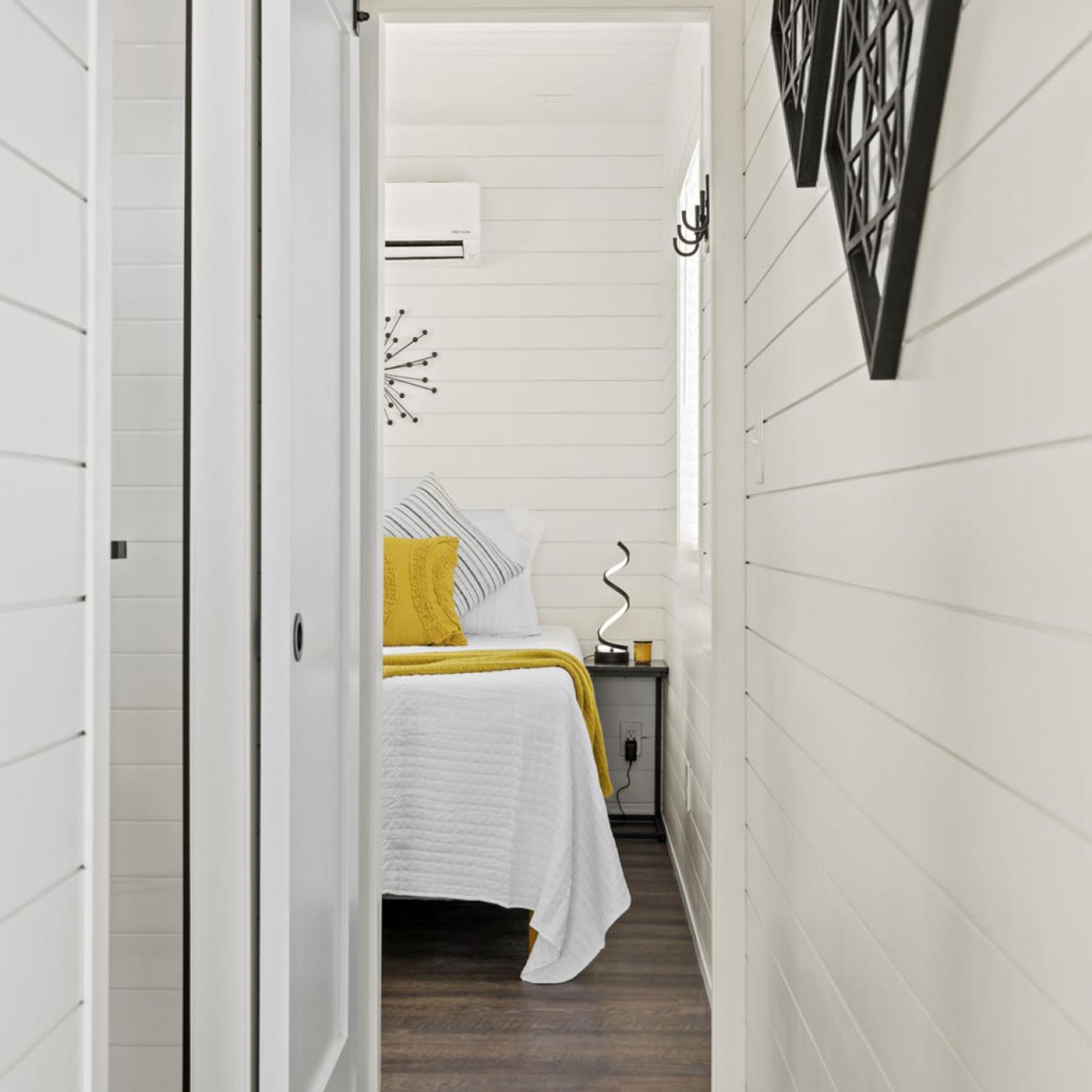 Een smalle gang die leidt naar een slaapkamer met een bed, een nachtkastje en een airconditioning.'