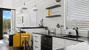 A modern kitchen with white cabinetry, black countertops, and a central island. There are hanging pendant lights, a built-in stove, and a window with blinds. - New The Yellow Beacon-luxury Shipping Container (Fredericksburg)