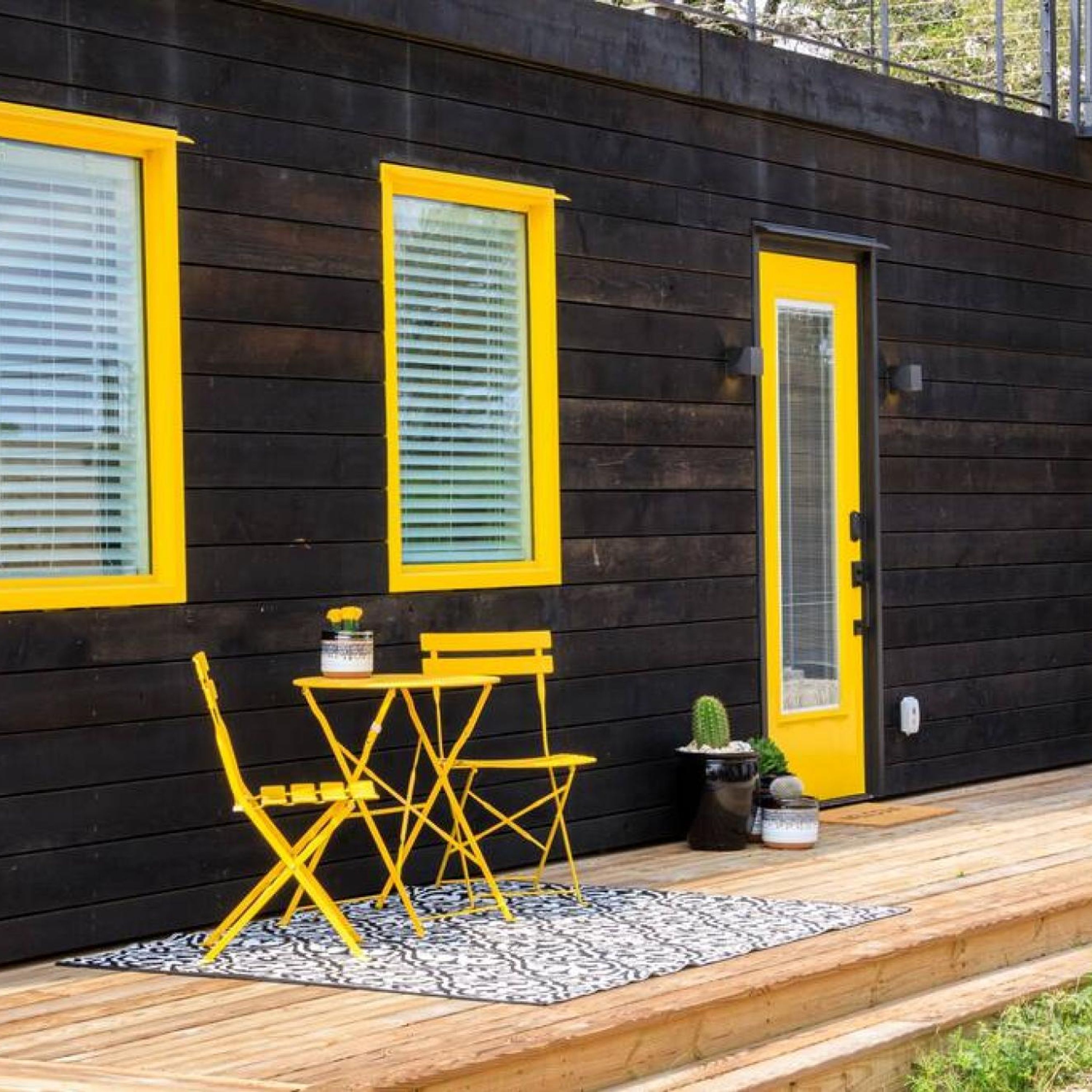 A wooden cabin with yellow-framed windows and a door, a small table with chairs, and a potted plant on the deck.