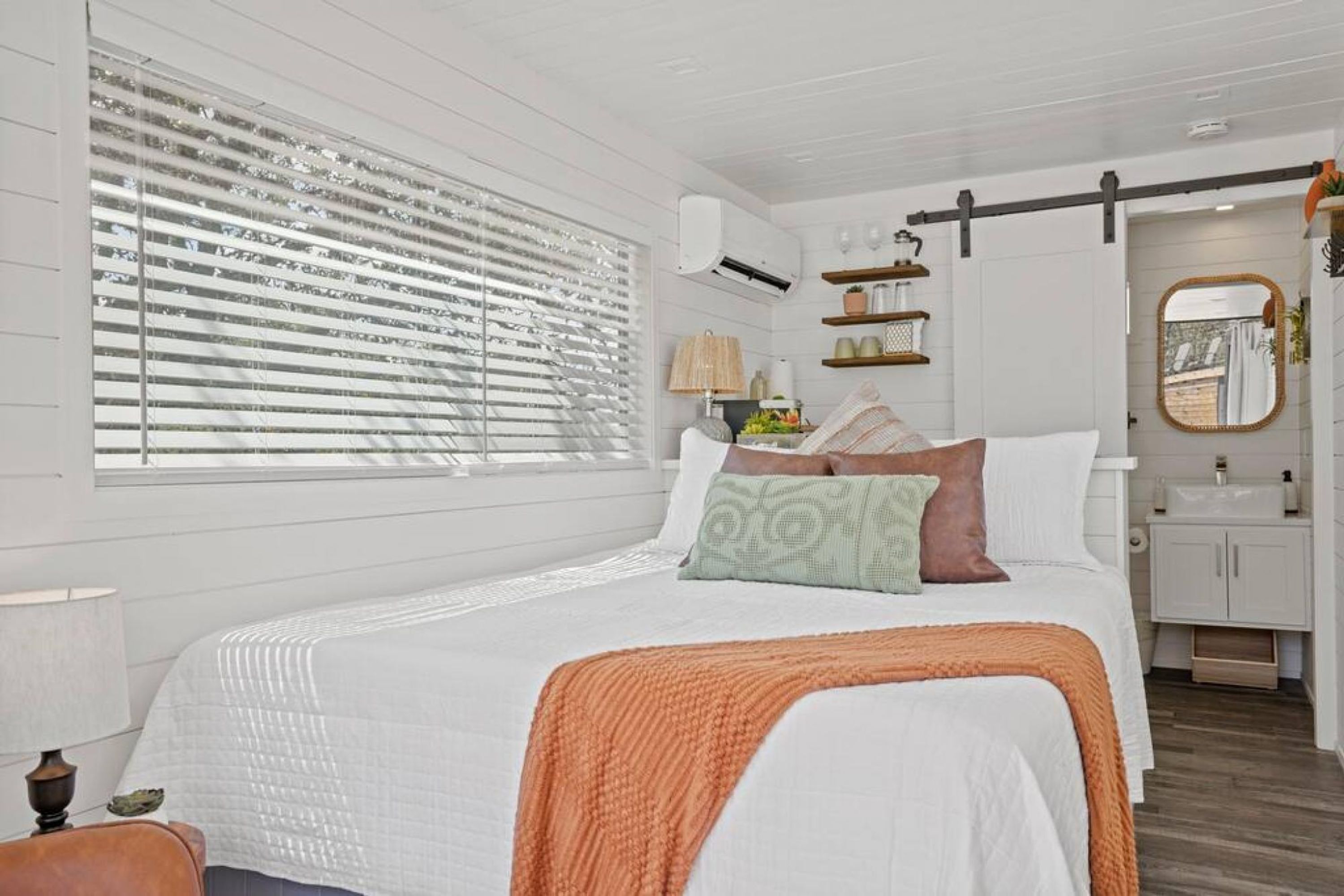Un dormitorio con cama, ventana con persianas, mesita de noche con lámpara y baño con lavamanos y espejo.