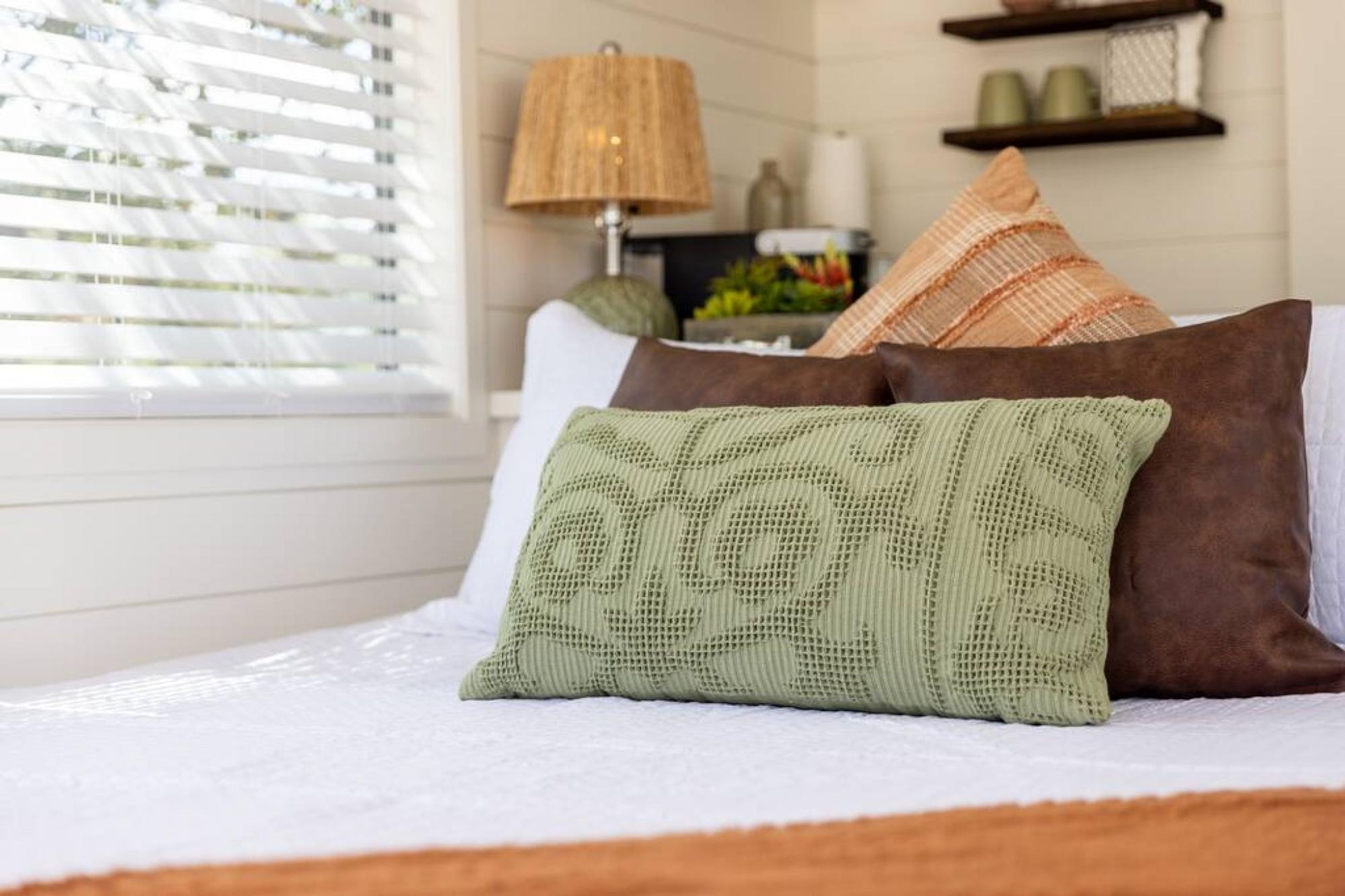A neatly made bed with a green knitted pillow and a brown pillow, a lamp, and a shelf with decorative items in the background.