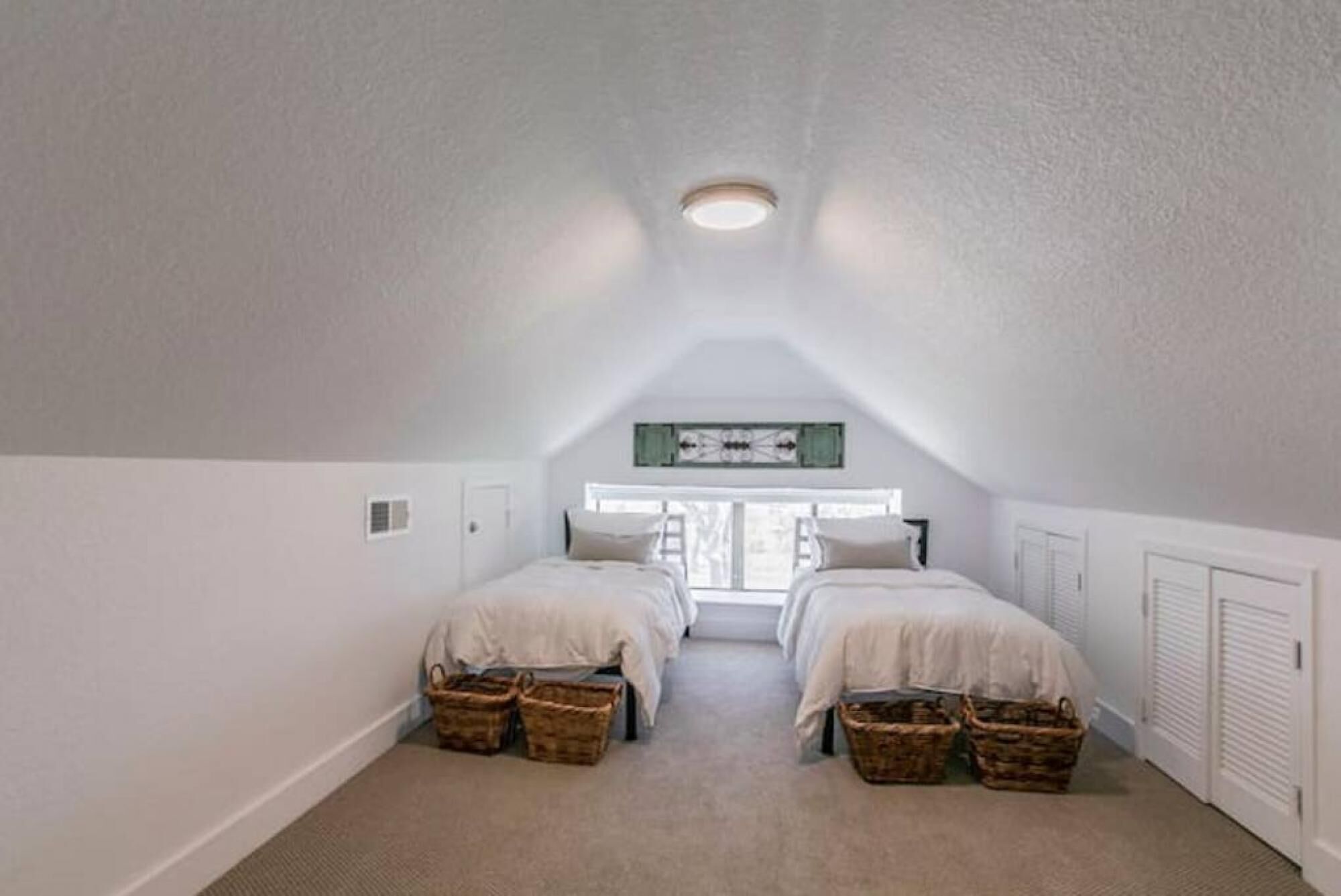 A small, well-lit attic bedroom with two beds, a sloped ceiling, and a large window.