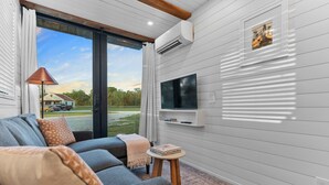 A modern living room with a grey sofa, a small wooden side table, a mounted TV, and a large window with curtains. - Cool River Helm Model Container Home 12 Minutes to Magnolia Baylor (Waco)