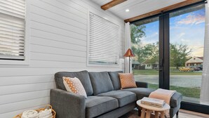 A modern living room with a grey sofa, a wicker basket, a small table, and a view of a residential street through a glass door. - Cool River Helm Model Container Home 12 Minutes to Magnolia Baylor (Waco)