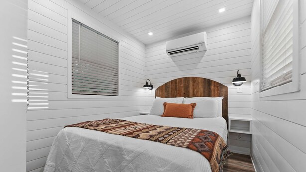 A bedroom with a wooden headboard, a white quilted bedspread, a patterned throw blanket, and a nightstand with a lamp. - Cool River Helm Model Container Home 12 Minutes to Magnolia Baylor (Waco)