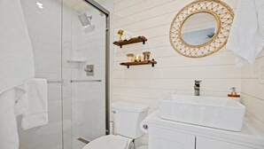 A bathroom with a glass shower enclosure, a white toilet, a white sink with a mirror above it, and a wooden shelf with jars. - New The Sunset Luxury Container Home (Fredericksburg)