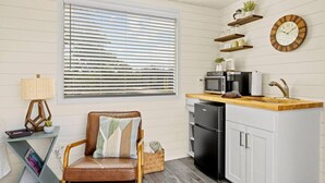 A compact kitchen with a refrigerator, microwave, and wooden countertop. There is a chair with a cushion and a small table with a lamp. - New The Sunset Luxury Container Home (Fredericksburg)