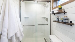 A modern bathroom with a glass-enclosed shower, white tiled walls, a toilet, and shelves with toiletries. - New The Lone Star Shipping Container (Fredericksburg)