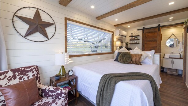 A bedroom with a large bed, a star decoration, a window with blinds, a wooden ceiling, a nightstand, and a dresser. - New The Lone Star Shipping Container (Fredericksburg)
