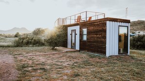 A small cabin with a wooden exterior and a metal roof, situated in a natural landscape with mountains in the background. - New Endless Sunsets-container Home (Alpine)