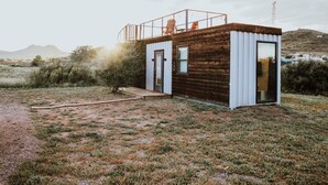 A modern cabin with a wooden exterior and a white door, situated in a grassy field with a clear sky. - New Endless Sunsets-container Home (Alpine)