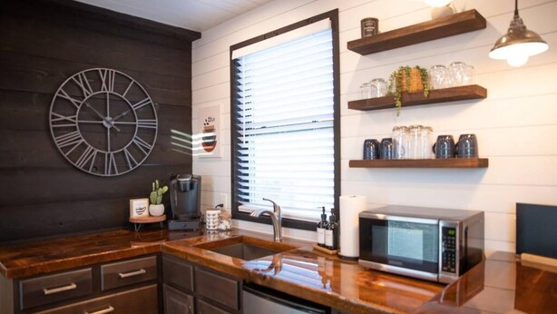 A modern kitchen with dark wooden cabinets, a stainless steel sink, a microwave, and a large window with blinds. - New Endless Sunsets-container Home (Alpine)