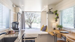 Cozinha compacta com cafeteira, pia e janela com persianas. Há uma cama com almofadas e uma luminária no lado direito.