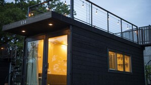 Uma cabana moderna com entrada por porta de vidro, varanda com luzes de cordão e janela visível com persianas.