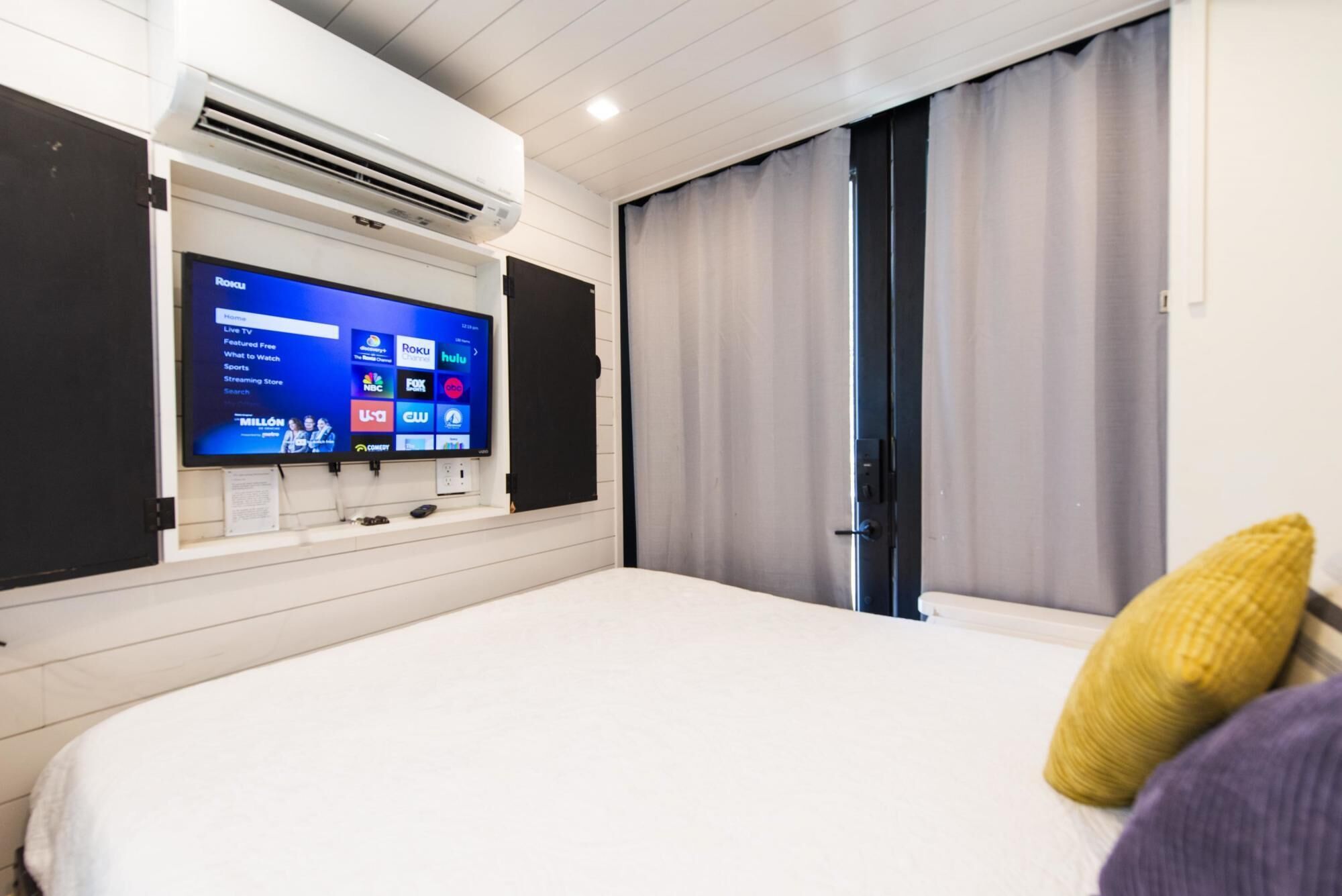 A hotel room with a bed, a mounted TV displaying various channels, and a window with curtains.