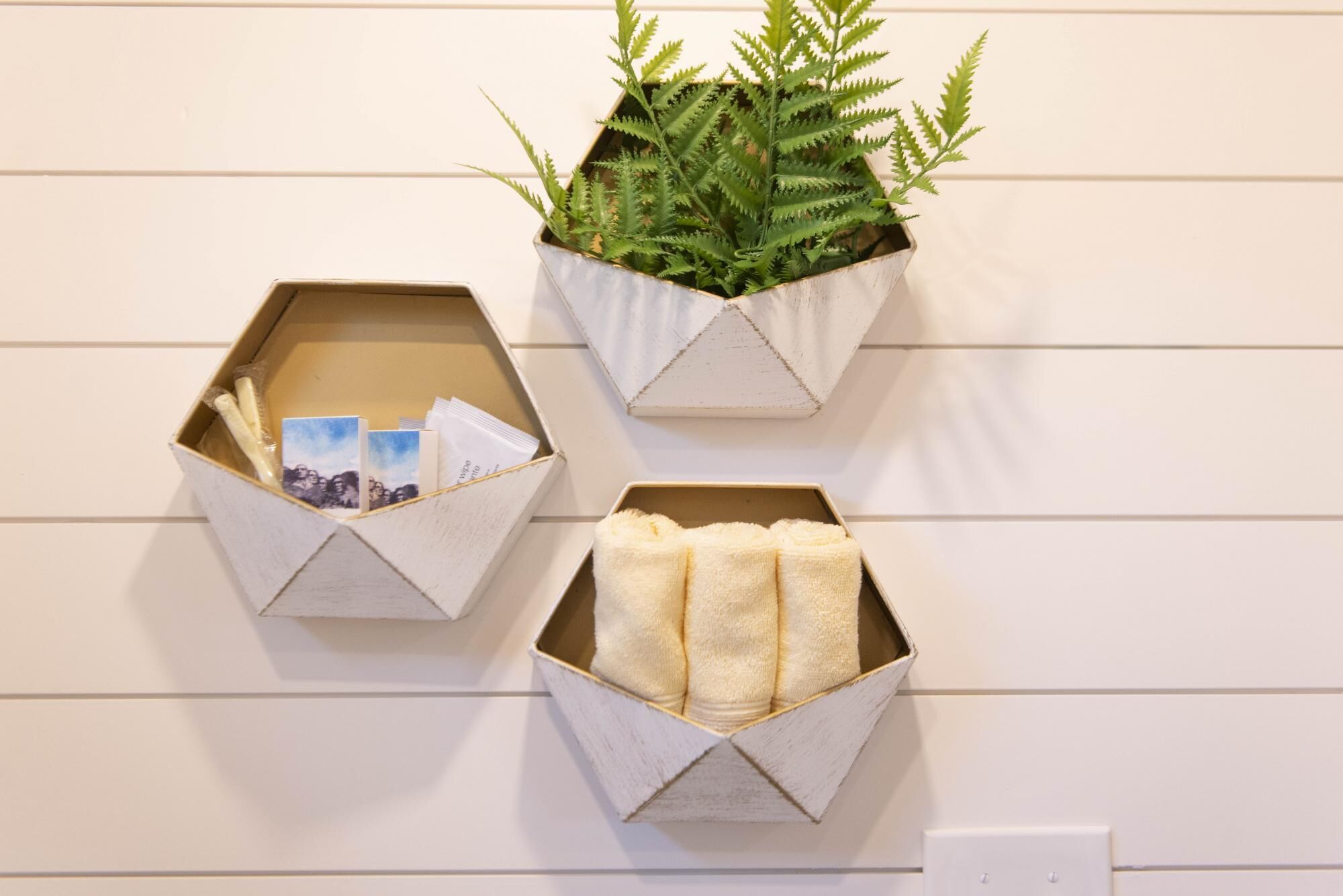Three hexagonal wall-mounted planters with different contents: one with a fern, one with cards, and one with towels.