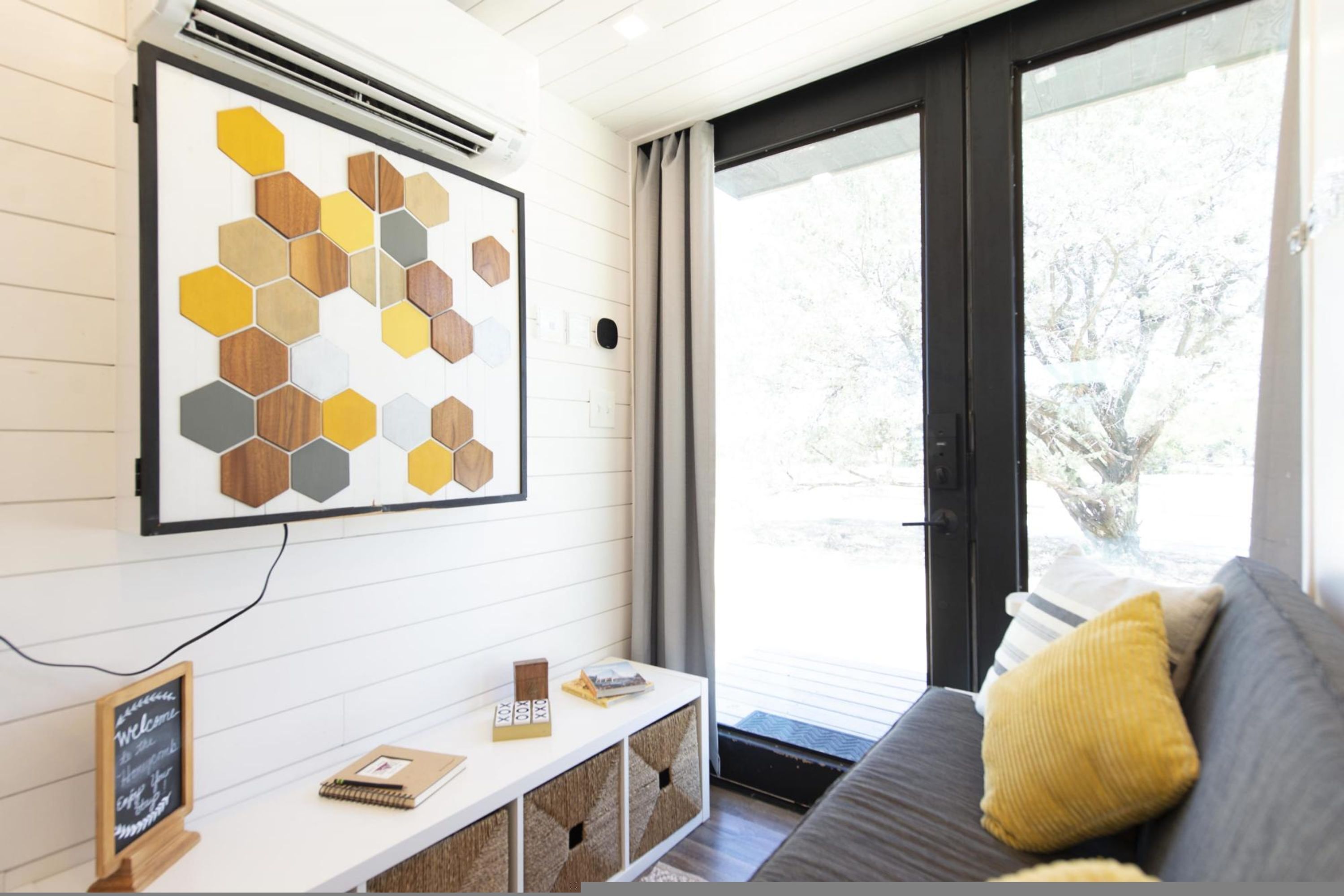 A modern living room with a white console table, a gray sofa with yellow cushions, and a large window with a view of trees.